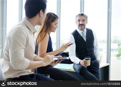 Smiling Asian team and Senior Caucasian boss is businesspeople teamwork relaxing and brainstorm. Businesswoman holding smartphone and talking about news at office. Focus at Caucasian man.