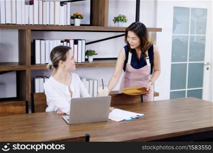 Smiling Asian waitress female serving a plasticcup of cold coffee for Caucasian businesswoman is relaxing with laptop on wooden desk at coffee shop.