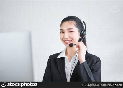 Smiling Asian woman consultant wearing microphone headset of customer support phone operator at workplace.