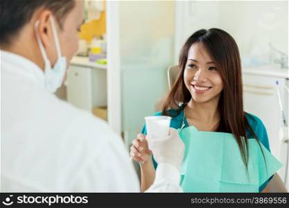 Smiling asian woman is taking a glass of water from her dentist