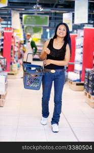 Smiling asian woman walking in grocery store carrying a shopping basket with store worker in the background