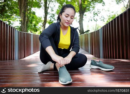 Smiling Asian young woman in black sportwear resting and tying Shoelace on wooden bridge before exercise and running in the garden.