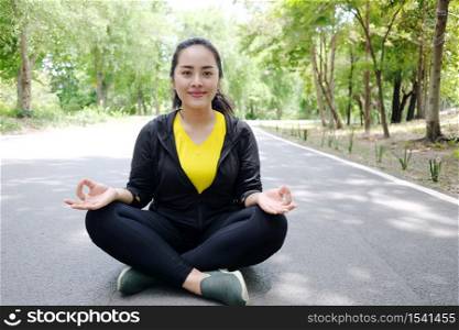 Smiling Asian young woman in sportwear sitting and doing yoga for meditation for create concentration and calm the mind on concrete floor after exercise in the natural garden.