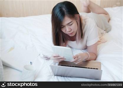 Smiling Asian young woman is laying on white bed holding tablet for online working and shopping with laptop in social medea in bedroom. Businesswoman work from home with Technology concept.