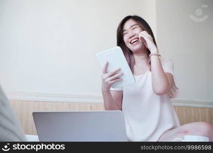 Smiling Asian young woman sitting on white bed is calling with smartphone and holding tablet for online working with laptop in bedroom. Businesswoman work from home with Technology concept.