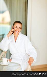 Smiling attractive female in bathrobe sitting at table on terrace and speaking phone&#xA;