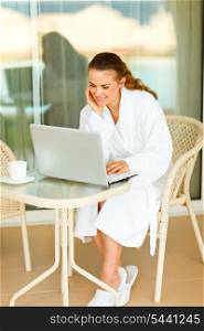 Smiling attractive female in bathrobe sitting at table on terrace and using laptop&#xA;