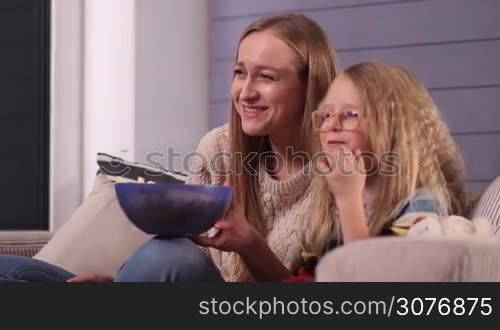 Smiling attractive mom and her lovely daugther in glasses sitting on cozy sofa in the living room and watching comedy show on tv and laughing while eating popcorn. Happy family enjoying watching television together in the evening.