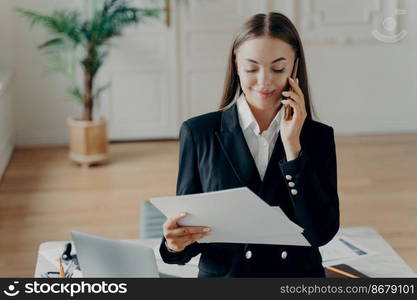 Smiling attractive young businesswoman in formal suit talking on phone and looking at notes, holding documents and discussing business ideas on smartphone, female manager standing in office. Successful young business lady talking on phone and looking at documents