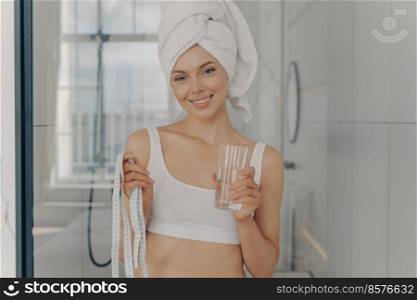 Smiling attractive young caucasian woman with glass of water in one hand and tape measure in other, standing in bathroom after morning shower, wears white underwear. Weightloss concept. Smiling attractive young caucasian woman with glass of water in one hand and tape measure in other