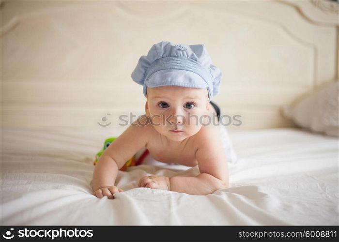 Smiling baby in blue cap playing with a toy car on the bed and looking at the camera. The baby on the bed