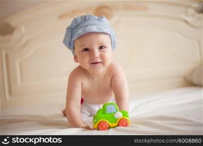 Smiling baby in blue cap playing with a toy car on the bed and looking at the camera. The baby on the bed