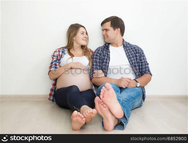 Smiling barefoot pregnant couple posing on floor at empty room