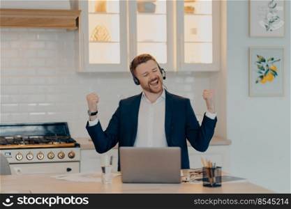 Smiling bearded businessman in suit and headset reaching business goals at work and celebrating success while sitting at table with laptop in kitchen, working remotely at home and enjoying victory. Happy manager in suit enjoying his work at home