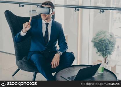 Smiling bearded businessman using modern VR headset glasses while sitting in office lobby and working on laptop, testing innovative technologies for managing business project through virtual reality. Smiling businessman using VR headset glasses while sitting in office lobby and working on laptop