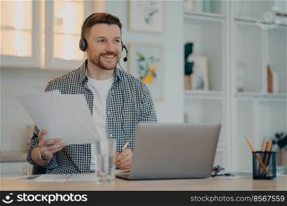 Smiling bearded guy sitting at his workplace and holding documents during online meeting, male freelancer working at home and using laptop, wearing headset. Remote job and freelance concept. Happy man holding papers during online call at home