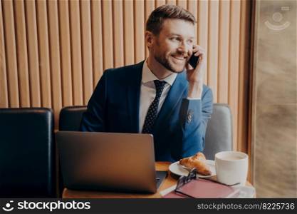 Smiling bearded handsome businessman in formal suit talking by cellphone while sitting with laptop at table in cafeteria having coffee break with cappuccino and croissant. Remote work indoor concept. Smiling unshaved handsome man of business in formal suit talking by cellphone in cafe