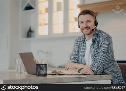 Smiling bearded handsome freelancer sitting at home and having video call while using headset and laptop, man smiling at camera during remote working day at home. Freelance concept. Cheerful guy enjoying freelance job while sitting at his workplace