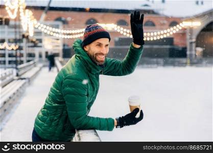 Smiling bearded male drinks takeaway coffee, waves with hand to his friend, has cheerful expression, going to have walk together. Outdoor shot of glad cheerful man in winter clothes with hot beverage