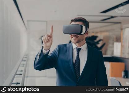 Smiling bearded office employee in suit wearing in VR glasses working in augmented reality in office, businessman in 3d goggles interacting with cyberspace at work, pointing with finger. Smiling bearded office employee in suit wearing in VR glasses working in augmented reality in office