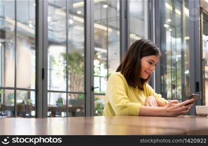 Smiling Beautiful Asian girl holding smartphone in coffee shop. Happy Pretty woman in yellow cloth sitting using smartphone while relaxing at cafe restaurant. lifestyle, copy space.