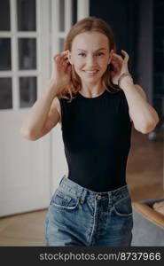 Smiling beautiful caucasian young woman fixing hairs at home, posing for photo portrait. Pretty female fashion model in casual clothes looking at camera with toothy smile, adjusting hair indoors.. Smiling pretty girl in casual clothes looking at camera adjusting hair posing for portrait indoors