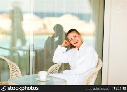 Smiling beautiful female in bathrobe sitting at table on terrace with cup of coffee&#xA;