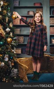 Smiling beautiful little girl stands on armchair, holds glass ball, decorates New Year tree, being happy to do it. Adorable small child busy with decoration of Christmas tree. Season and celebration