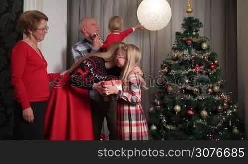 Smiling beautiful mother giving Christmas wrapped gift box to little daugther and kissing her with tenderness. Excited girl opening gift box and giving kiss to grandmother. Happy family with grandparents giving presents to children by Christmas tree.