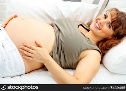Smiling beautiful pregnant female lying on sofa at home and holding her belly.&#xA;