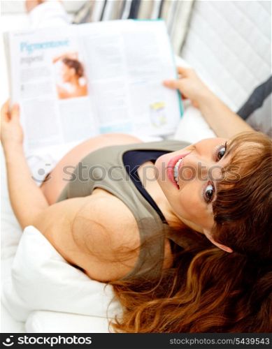 Smiling beautiful pregnant female reading magazine while sitting on sofa at home.&#xA;