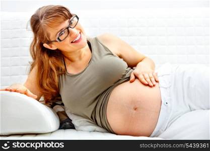 Smiling beautiful pregnant female relaxing on sofa at home and holding her belly.&#xA;