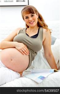 Smiling beautiful pregnant female relaxing on sofa at home with magazine.&#xA;