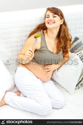 Smiling beautiful pregnant female sitting on divan at home and eating pickle&#xA;