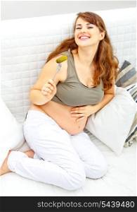 Smiling beautiful pregnant female sitting on divan at home and eating pickle&#xA;