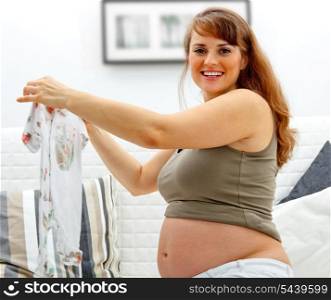 Smiling beautiful pregnant female sitting on sofa at home with baby clothes in hands.&#xA;