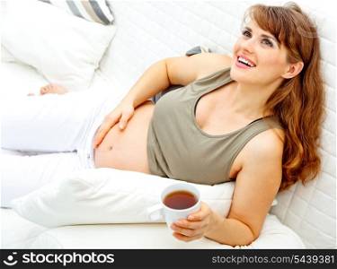 Smiling beautiful pregnant woman relaxing on couch at home and holding cup of tea in hand&#xA;