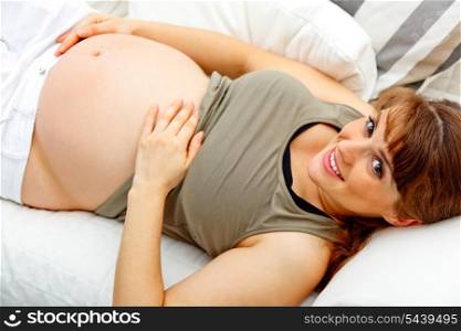 Smiling beautiful pregnant woman relaxing on couch at home and holding her belly&#xA;