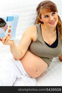 Smiling beautiful pregnant woman relaxing on couch at home with magazine.&#xA;