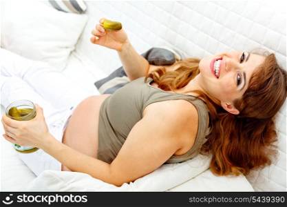 Smiling beautiful pregnant woman relaxing on sofa at home and holding jar of pickles in hands&#xA;