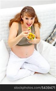 Smiling beautiful pregnant woman sitting on couch at home and eating fruit salad&#xA;