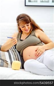 Smiling beautiful pregnant woman sitting on sofa and knitting for her baby.&#xA;