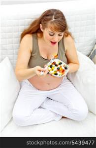 Smiling beautiful pregnant woman sitting on sofa at home and eating fresh vegetable salad&#xA;