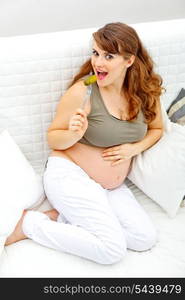 Smiling beautiful pregnant woman sitting on sofa at home and eating pickle&#xA;