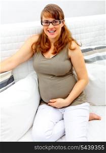 Smiling beautiful pregnant woman sitting on sofa at home and holding her belly.&#xA;