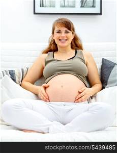 Smiling beautiful pregnant woman sitting on sofa at home and touching her belly.&#xA;