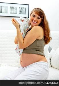 Smiling beautiful pregnant woman sitting on sofa at home with baby clothes in hands.&#xA;