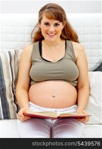Smiling beautiful pregnant woman sitting on sofa at home with book&#xA;