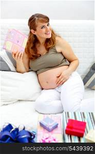 Smiling beautiful pregnant woman sitting on sofa at home with gifts for her unborn baby&#xA;