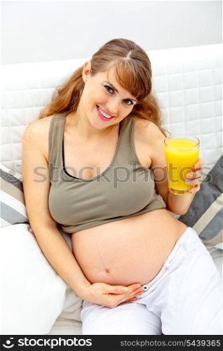 Smiling beautiful pregnant woman sitting on sofa at home with glass of juice in hand&#xA;